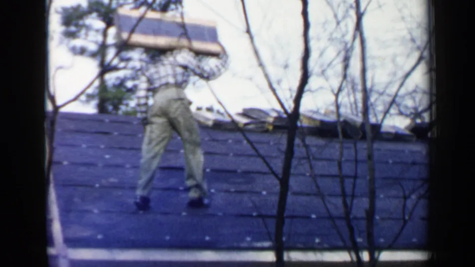 A man crosses the sloped roof with roofing material, seen through bare branches in the 1957 Birmingham home-movie footage.