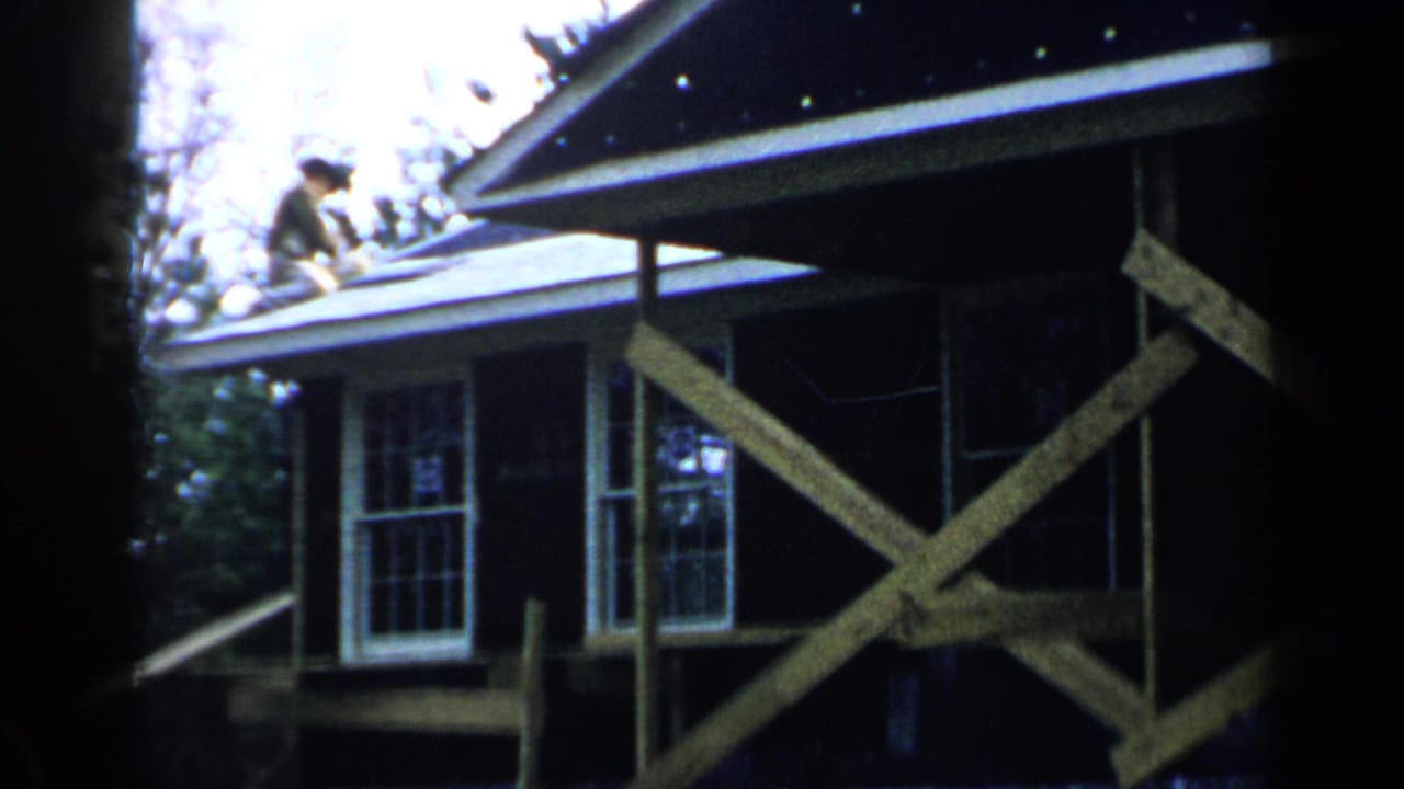 Man working on a house roof beside wooden scaffolding and white-trimmed windows.