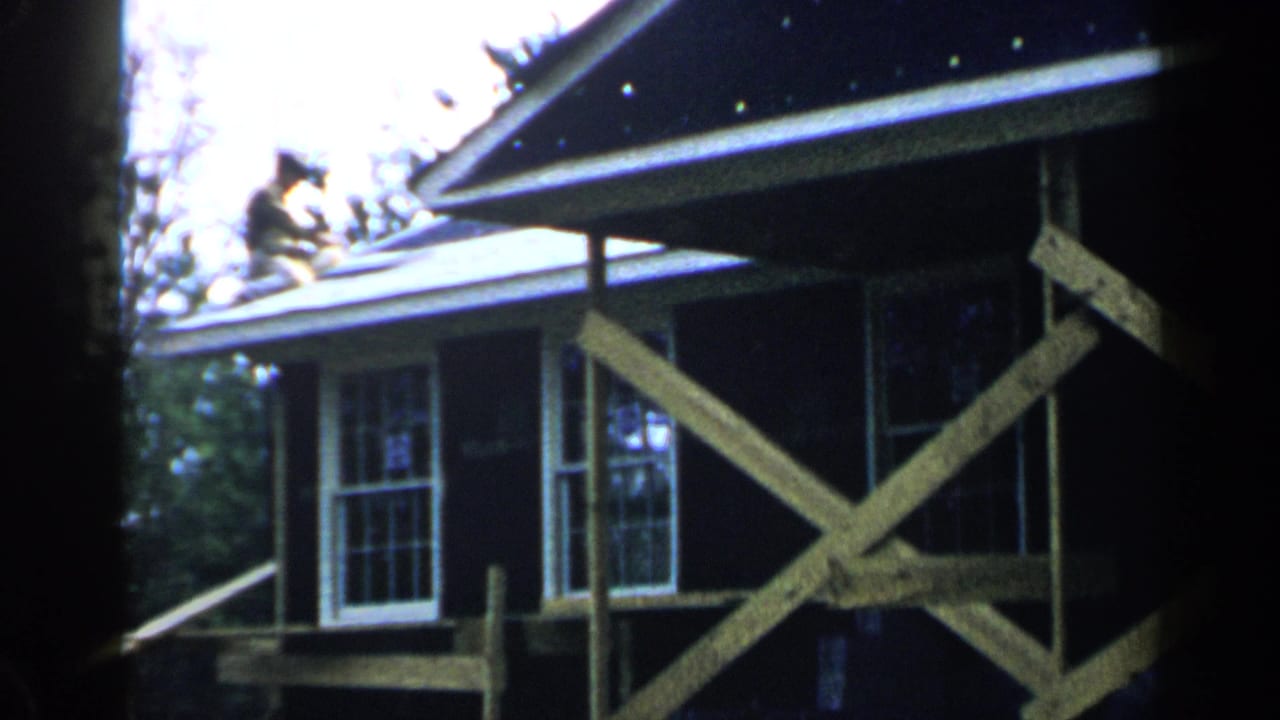 Man on roof near the edge of a dark-sided house with cross-braced scaffolding.