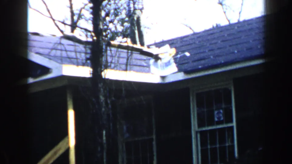 1957 Birmingham home-movie view of a tree trunk before a dark building with roof eaves, window, and exposed lumber.