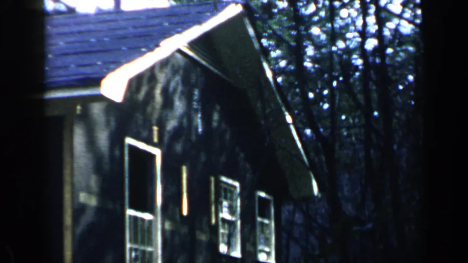 1957 Birmingham home-movie frame showing a dark-sided house in woods, with roof edge, windows, and strong daylight contrast.