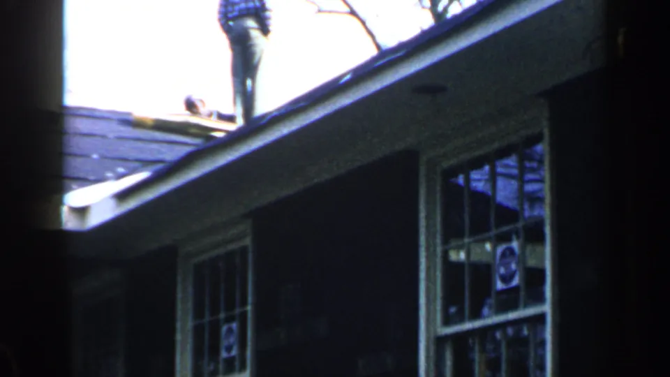 1957 Birmingham home-movie frame with a man on a sloped roof above multi-pane windows on a wooded house exterior.