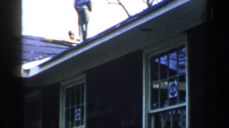1957 Birmingham frame showing a man standing near the roof edge above a dark-sided house with multi-pane windows.