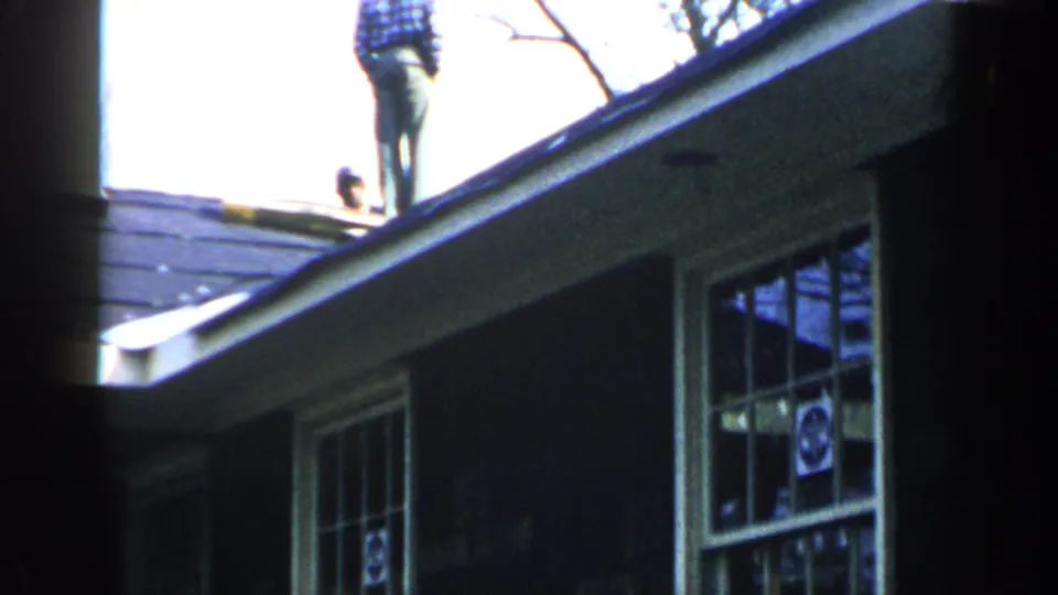 1957 Birmingham home-movie frame showing a man on a roof edge, another partial figure, dark shingles, white eaves, and windows.