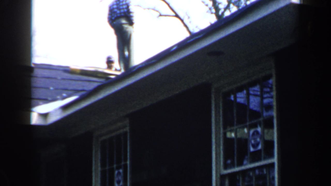 Man stands near the roof peak above a house with windows and eaves.