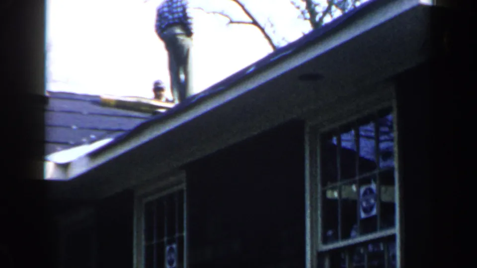 A man in a plaid shirt stands near the roof peak of a Birmingham house, with windows, eaves, and bright sky visible.