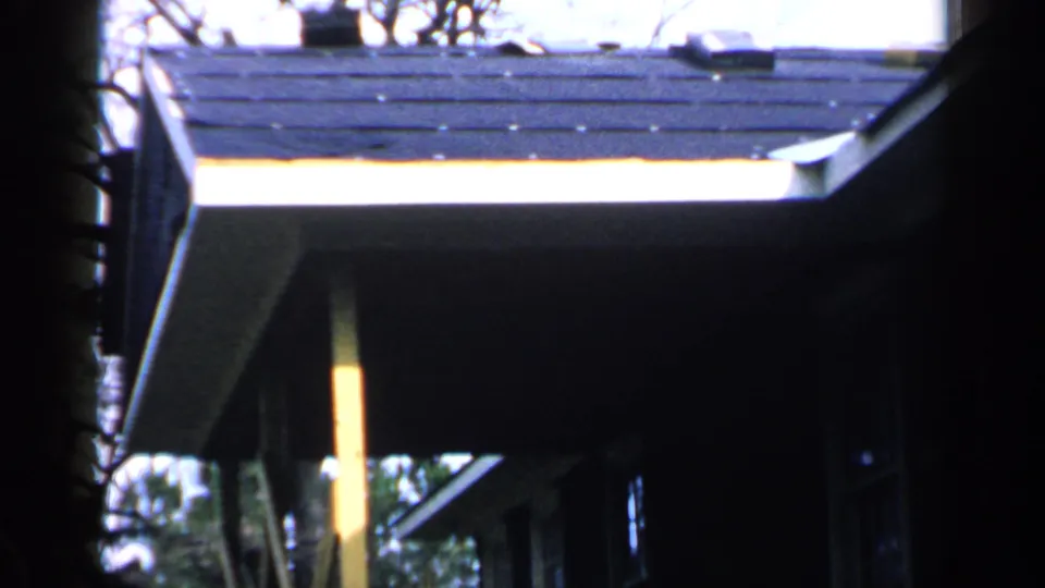 1957 Birmingham 8mm frame showing a house roofline with dark shingles, overhanging eaves, a yellow post, and winter-like trees.
