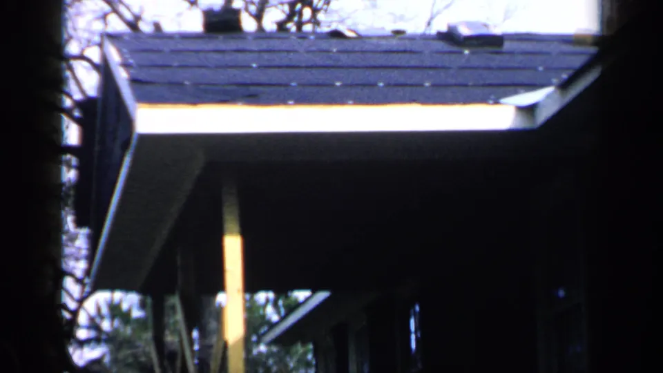 1957 Birmingham home-movie frame of a shadowed house roofline, bright fascia light, bare branches, and a yellow support post.