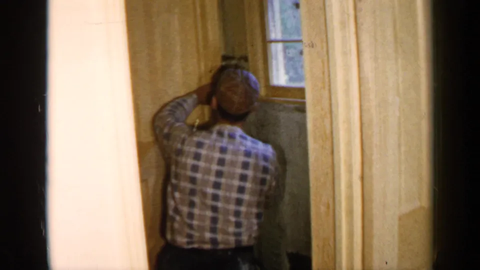 1957 Birmingham home repair frame showing a man in a plaid shirt and cap working on wood paneling beside a window.