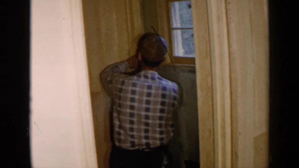 1957 Birmingham frame of a man seen from behind during interior home repair near a window and unfinished wood paneling.