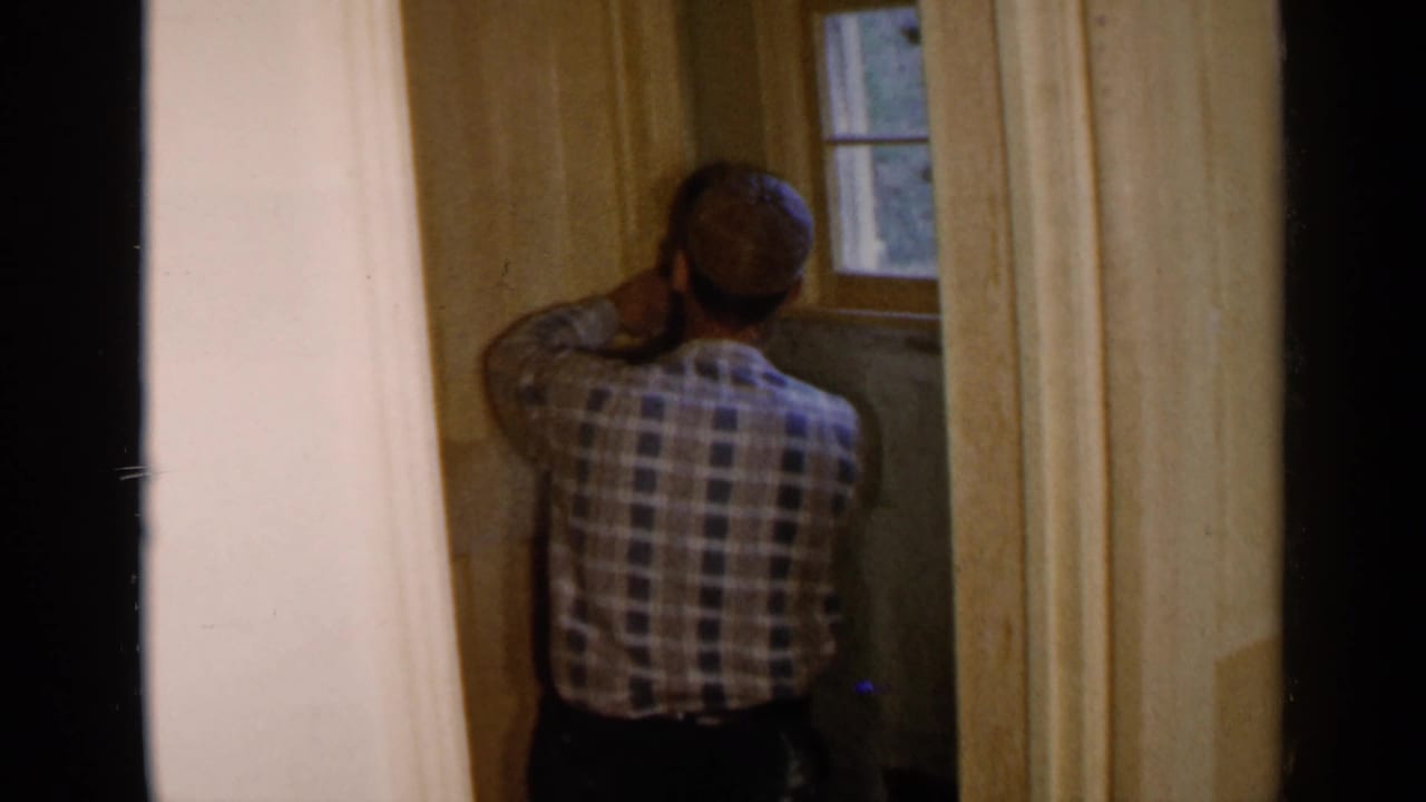 Man in plaid shirt and cap working on an unfinished wall beside a small window.