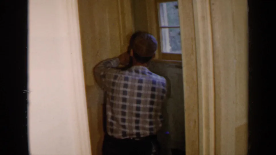 In 1957 Birmingham home-movie footage, a man in a plaid shirt works on an unfinished interior wall near a small window.