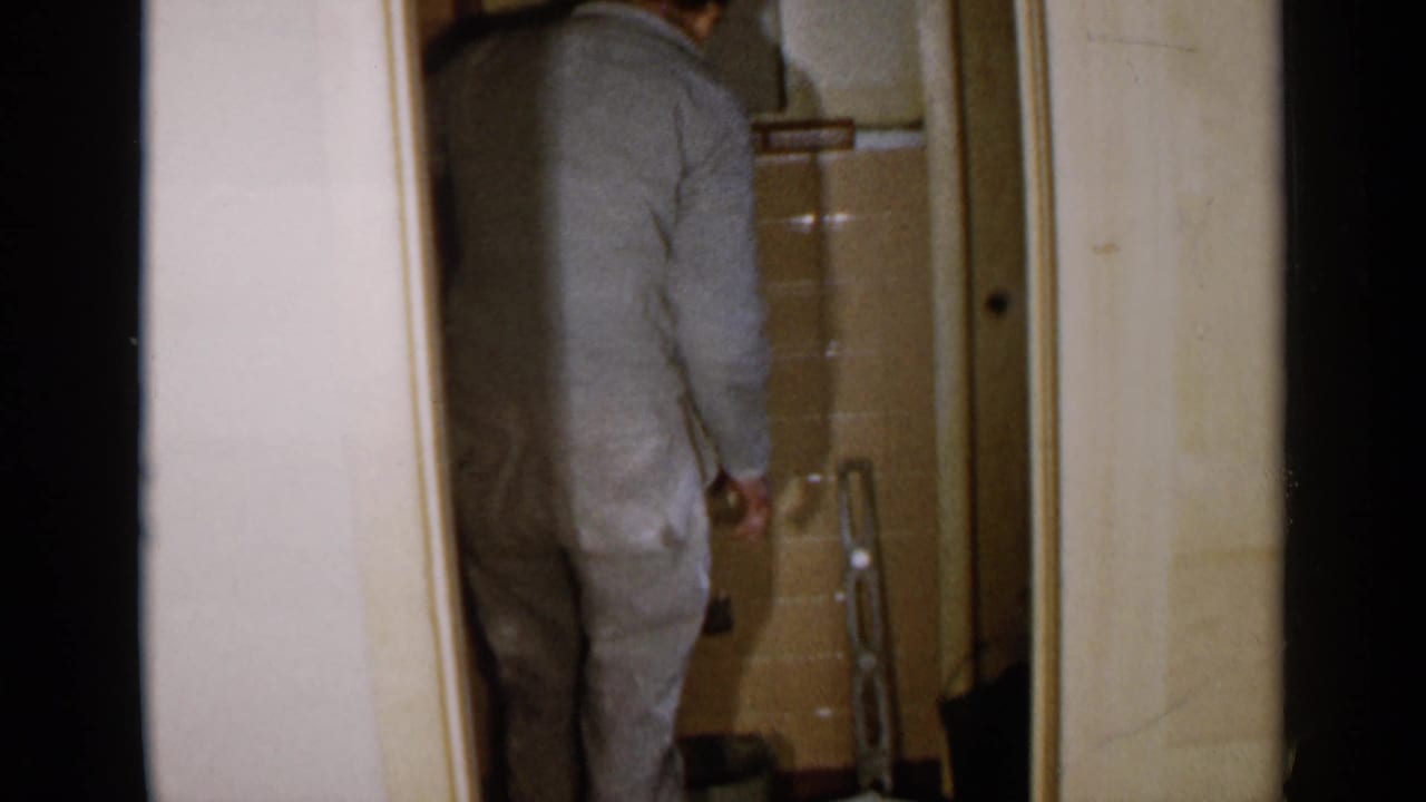 Man in a gray jumpsuit standing in a tiled bathroom doorway near plumbing fixtures.