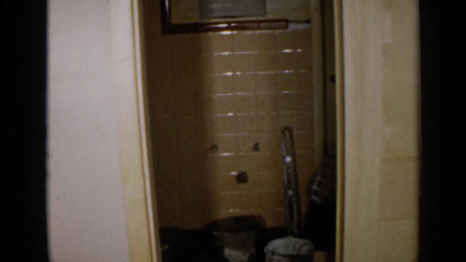 The bathroom is mostly empty after the worker moves out of view, with beige tile, pipes, and floor items visible in the doorway.