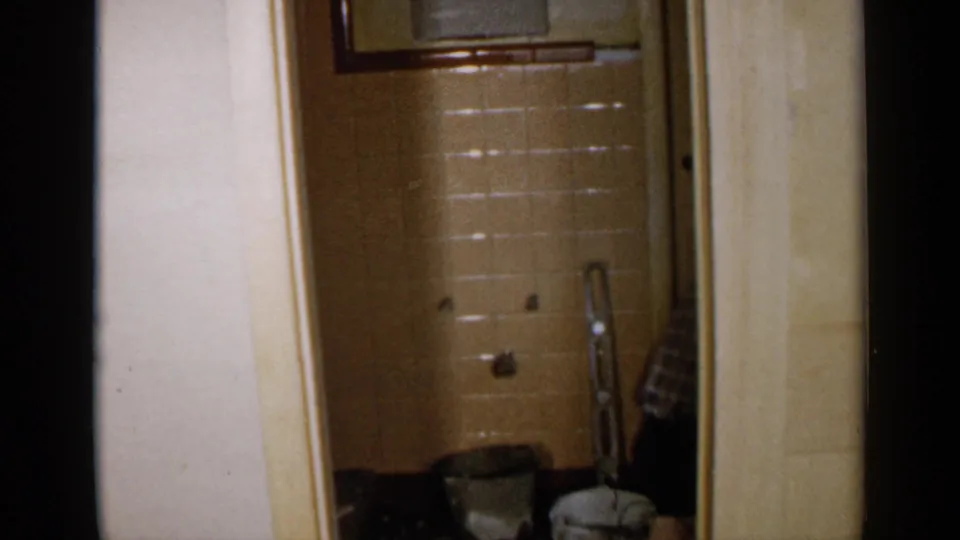 A 1957 Birmingham bathroom interior shows beige tile, plumbing fixtures, a small ladder, and supplies after the worker has stepped away.