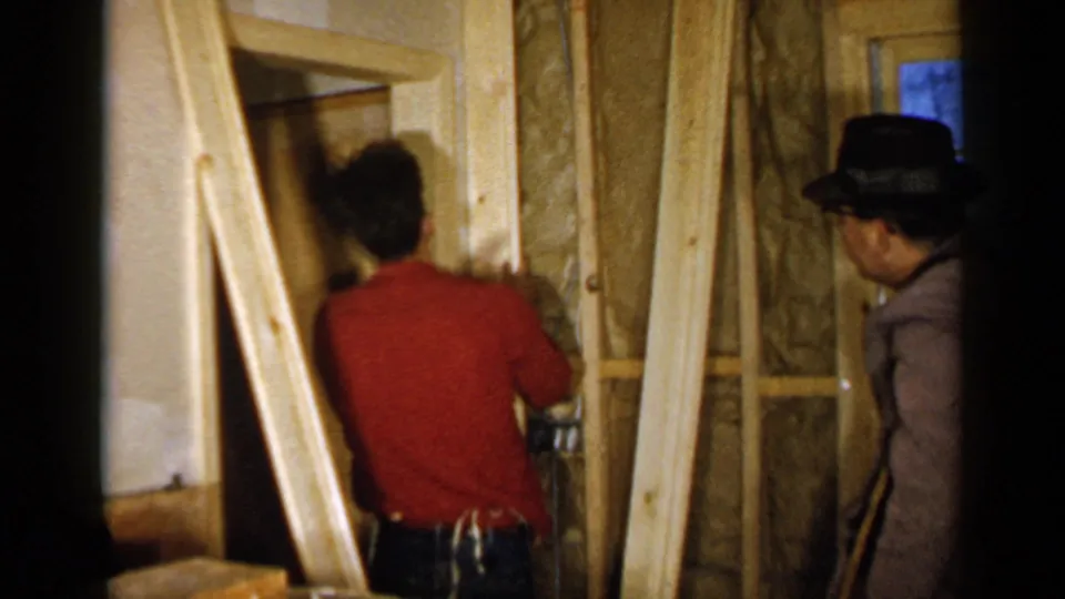 1957 Birmingham home renovation scene with a man in a red shirt working at exposed framing as a second man observes.