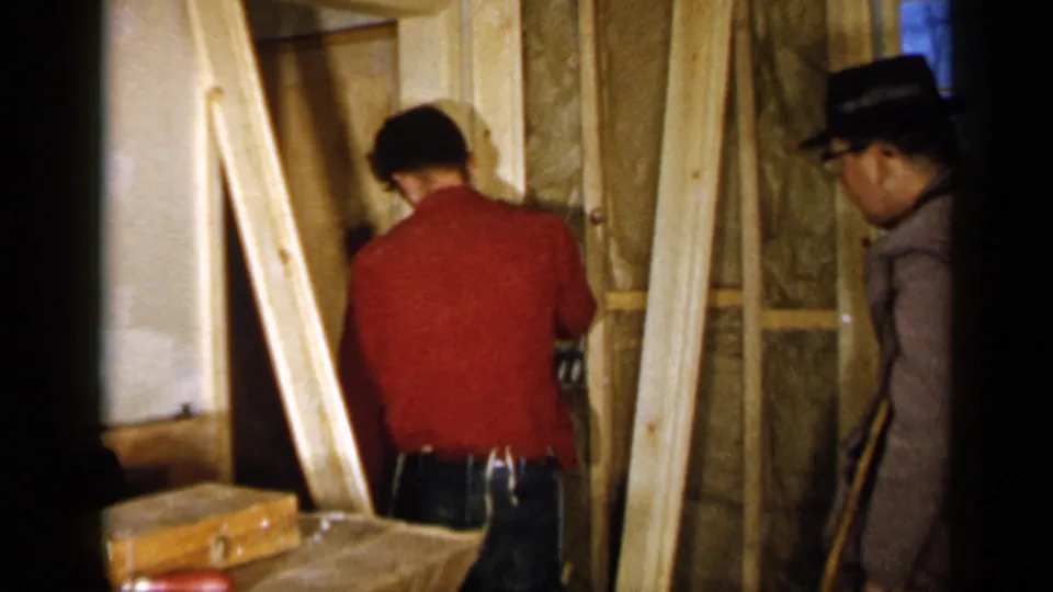 A man in a red shirt works on wood framing inside a Birmingham residence, with exposed insulation and another adult nearby.