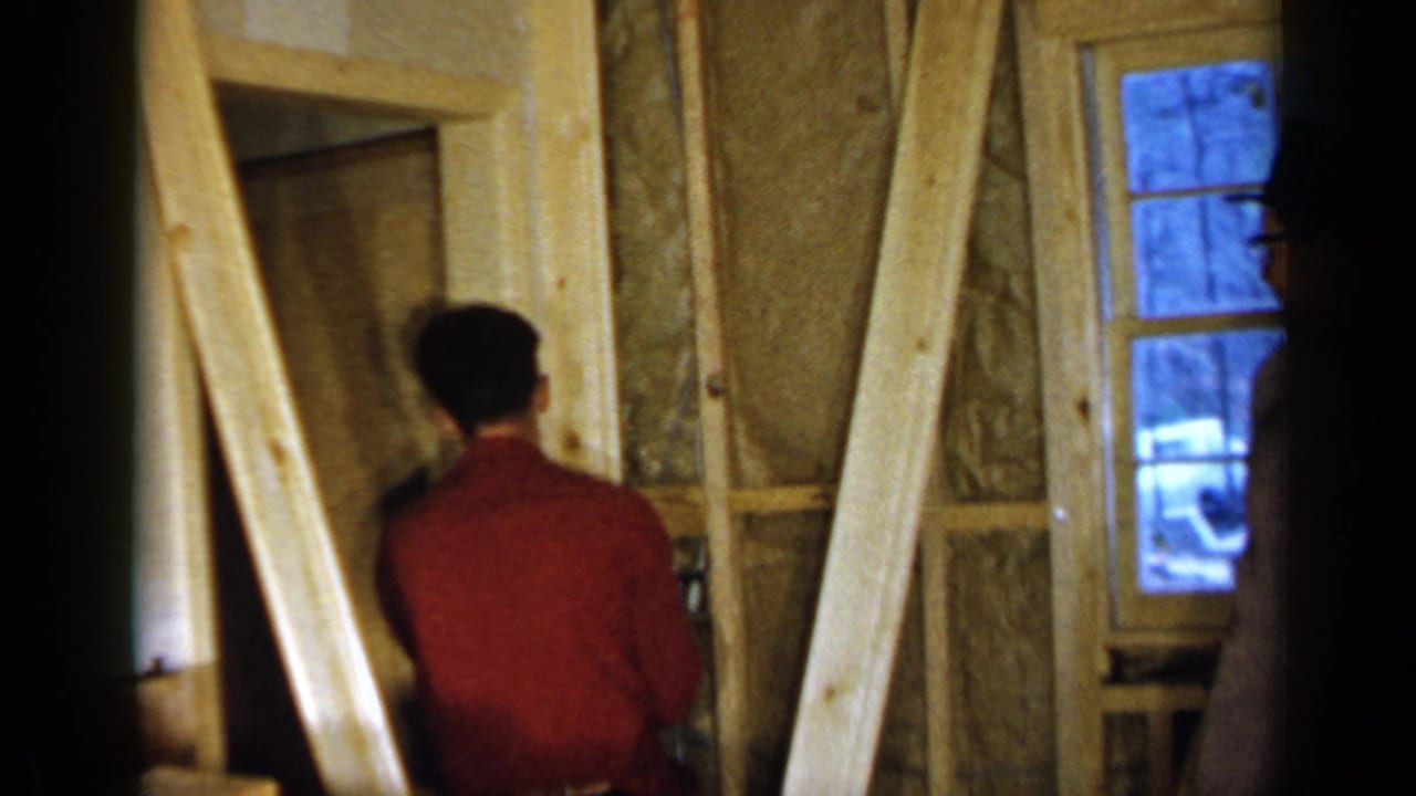 Man in a red shirt works beside exposed studs and insulation inside a wooden structure.