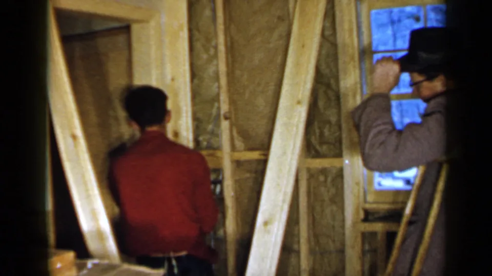 Two adult men work inside a partially constructed Birmingham interior, with a window, exposed studs, insulation, and loose lumber visible.