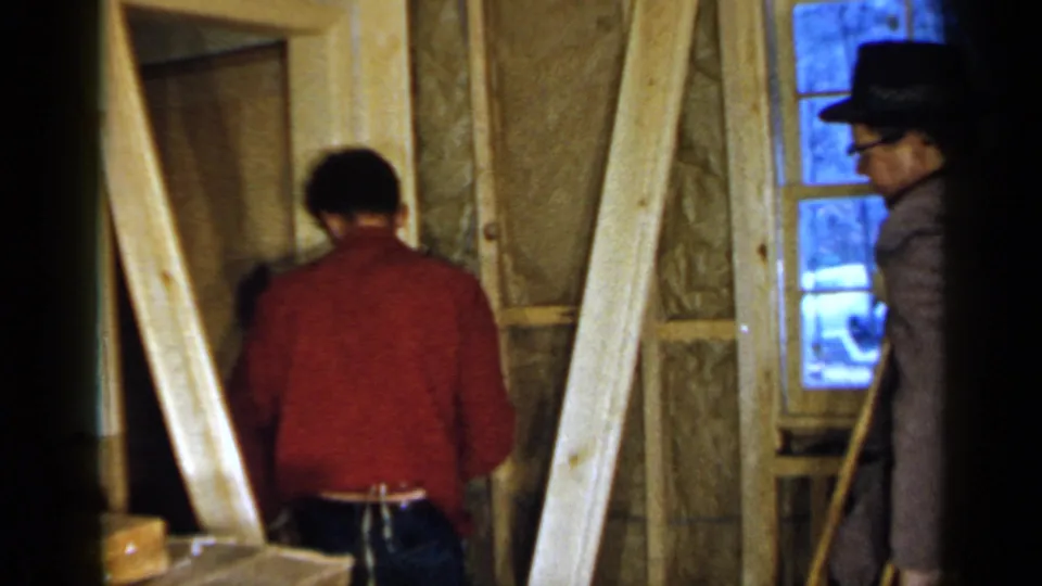A man in a red shirt works close to exposed framing while a second man in a dark hat and glasses observes near the window.