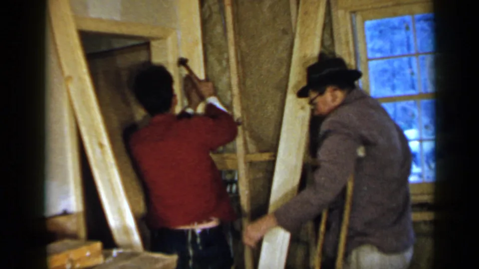 Inside the 1957 Birmingham construction scene, one man raises a hammer near the framing while another man in a hat assists nearby.