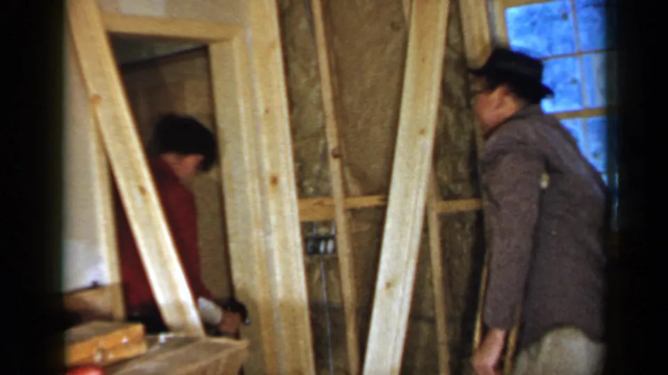 The red-shirted man leans toward the doorway frame while the hat-wearing man with crutches observes in the unfinished Birmingham interior.