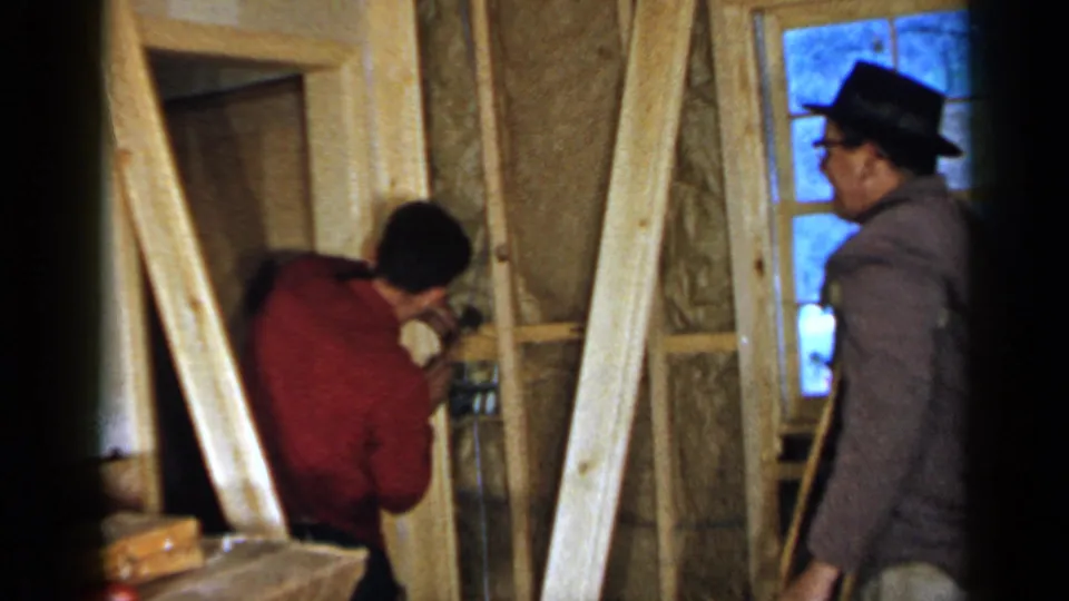 Inside the unfinished room, the red-shirted man works close to the frame as the man in a hat stands to the side with crutches visible.