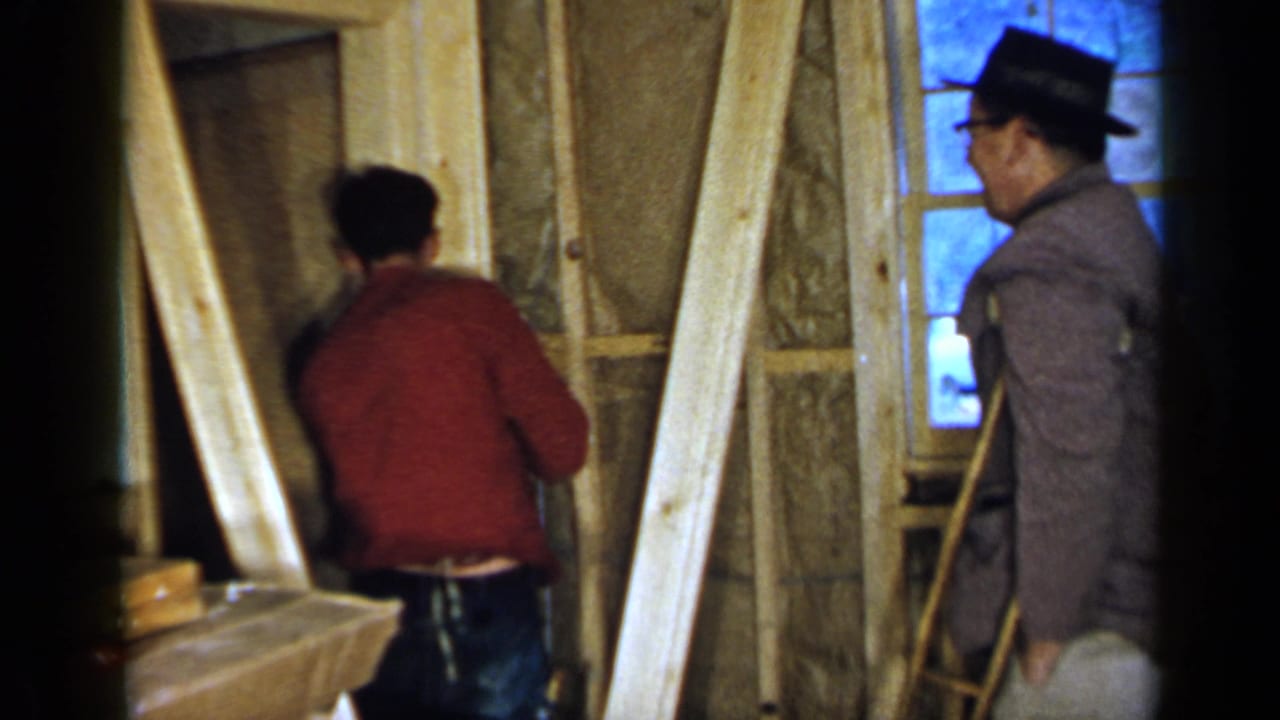 Man in a red sweater hammers an unfinished interior wall while another man in a hat stands nearby.
