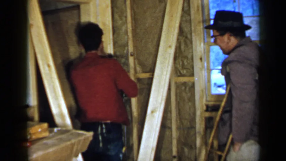 Two men appear in a 1957 Birmingham interior construction scene, with one facing the wall and the other observing near a window and lumber.