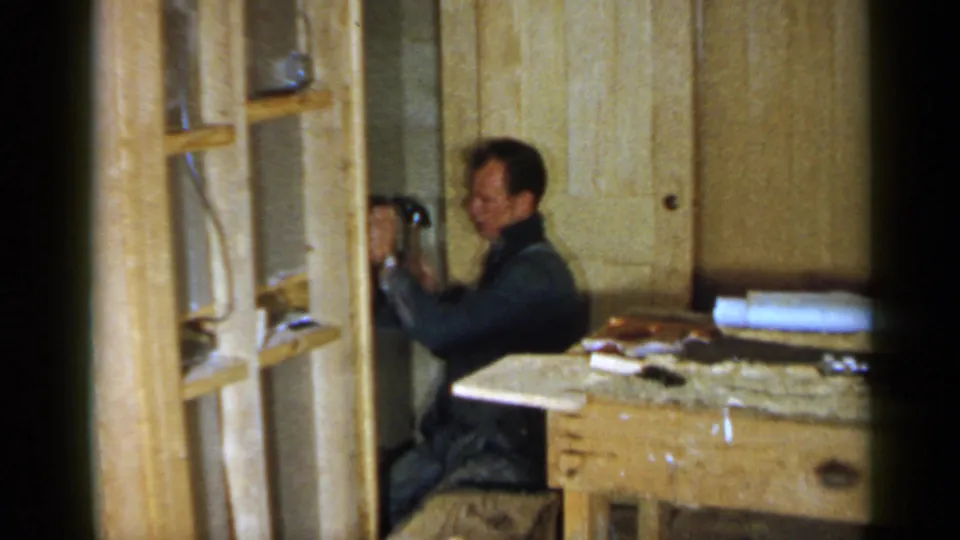 A man works inside a 1957 Birmingham unfinished interior near a door, shelving, workbench, and scattered building materials.