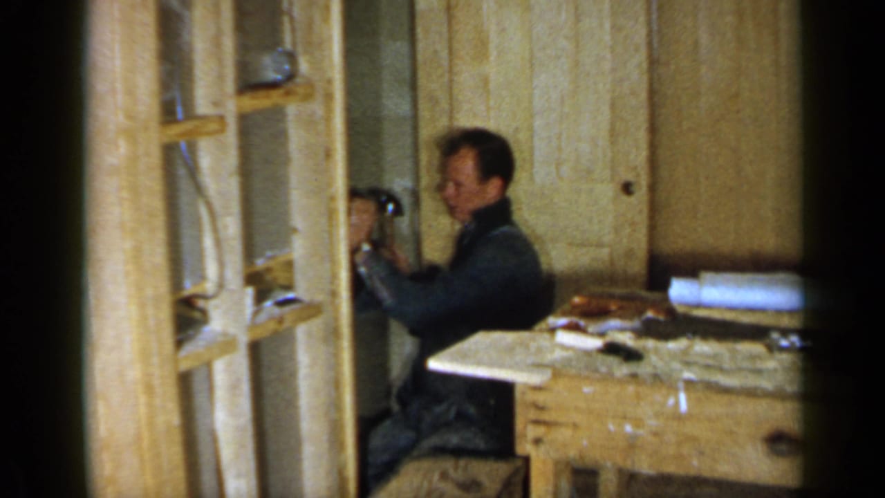 In 1957 Birmingham home-movie footage, a man works with a hammer beside exposed wood framing and a cluttered bench.