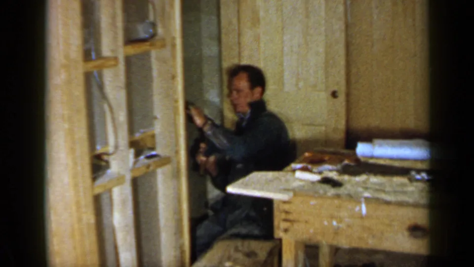 A man continues a small woodworking or repair task beside exposed framing in the Birmingham home-movie scene.
