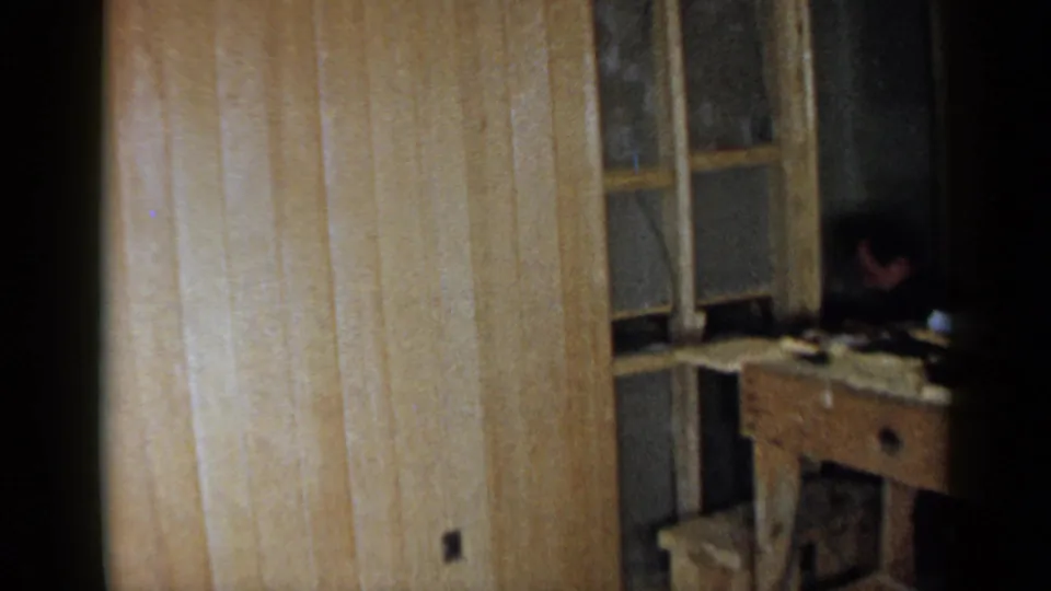 1957 Birmingham home renovation frame showing paneling, window framing, a rough wooden workbench, a stool, and a person partly visible in shadow.