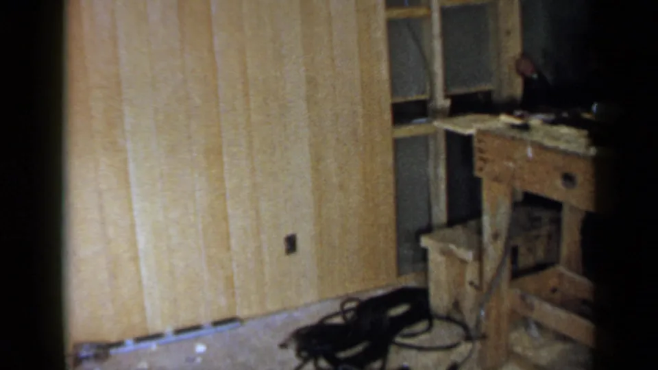 1957 Birmingham interior repair view with pale wall paneling, window framing, a workbench, a stool, scattered materials, and a dark floor object.