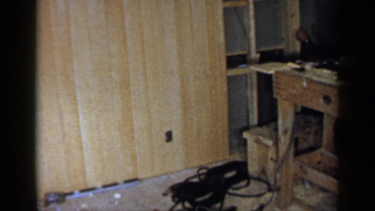 Wood-paneled room with a workbench, chair, floor debris, and dark film edge.