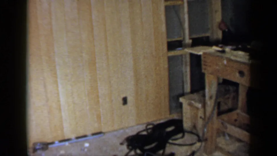 1957 Birmingham home-movie frame showing a wood-paneled interior with a workbench, chair, scattered floor debris, and visible film framing.