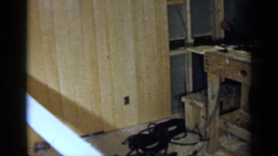 1957 Birmingham home-movie frame of a private wood-paneled room with a workbench, chair, floor debris, and bright diagonal film flare.