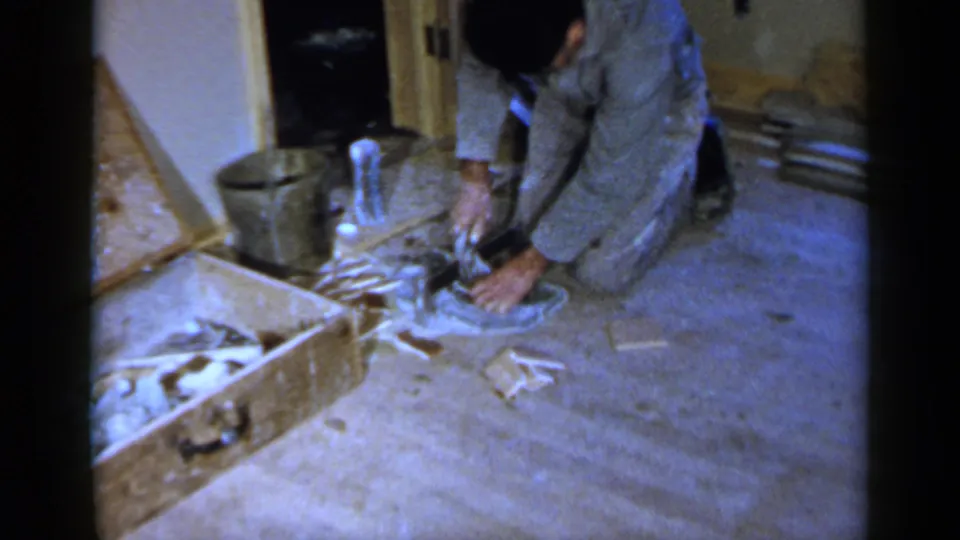 1957 Birmingham home-movie frame showing a man in a grey shirt kneeling on carpet and using a small silver tool near a wooden box.