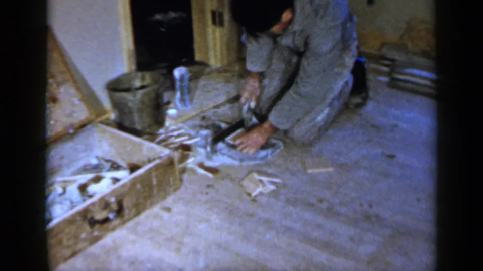 1957 Birmingham home-movie frame of a man kneeling in a private room, working with a small tool beside cut scraps and a wooden box.