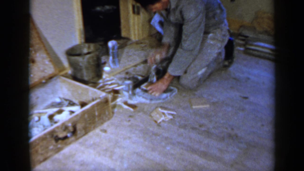 Man in gray work clothes kneels on a floor beside tools and renovation debris.