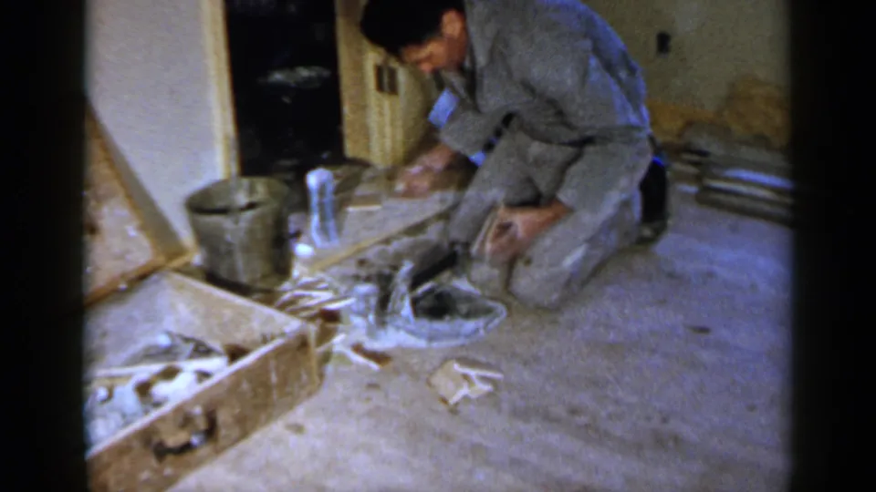 The home-movie frame shows a kneeling man reaching across a work area during a 1957 residential floor repair scene.