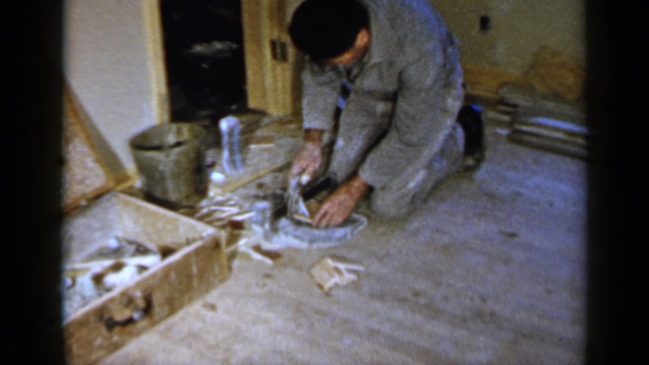Man in gray work clothes kneels on a concrete floor beside a crate and bucket.