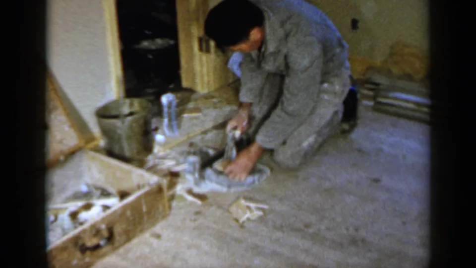 A close home-movie view shows the worker reaching down in a cluttered unfinished room in Birmingham, Alabama.