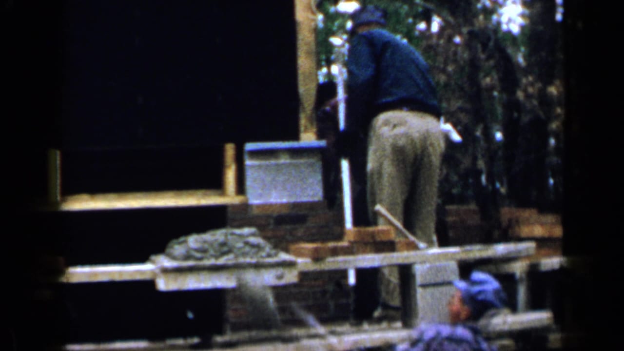 Worker on a wooden scaffold beside brickwork and mortar at an outdoor construction site.