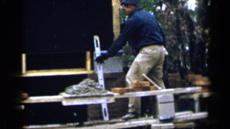A Birmingham construction worker reaches beside a ladder on the scaffold, with mortar and bricks visible in the foreground.