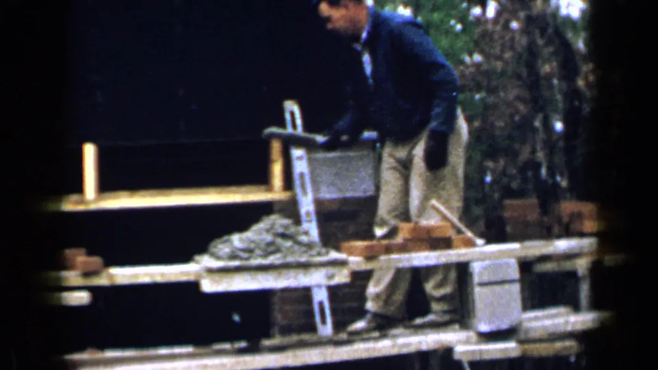 A worker on the scaffold stands near the mortar and ladder while another worker in a cap is partly visible below.