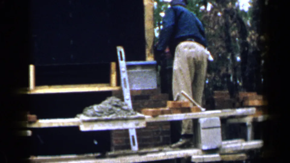 The scaffold-level worker faces the wall in a 1957 Birmingham masonry scene, with mortar, bricks, and ladder in view.