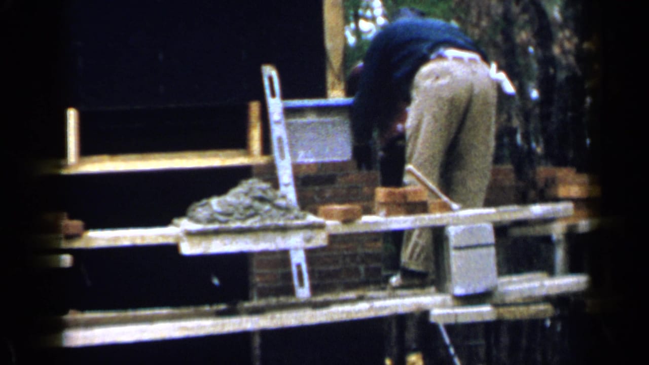 In 1957 Birmingham footage, a construction worker bends on scaffold planks near bricks, mortar, and a partially built wall.