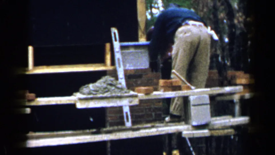 In 1957 Birmingham footage, a construction worker bends on scaffold planks near bricks, mortar, and a partially built wall.