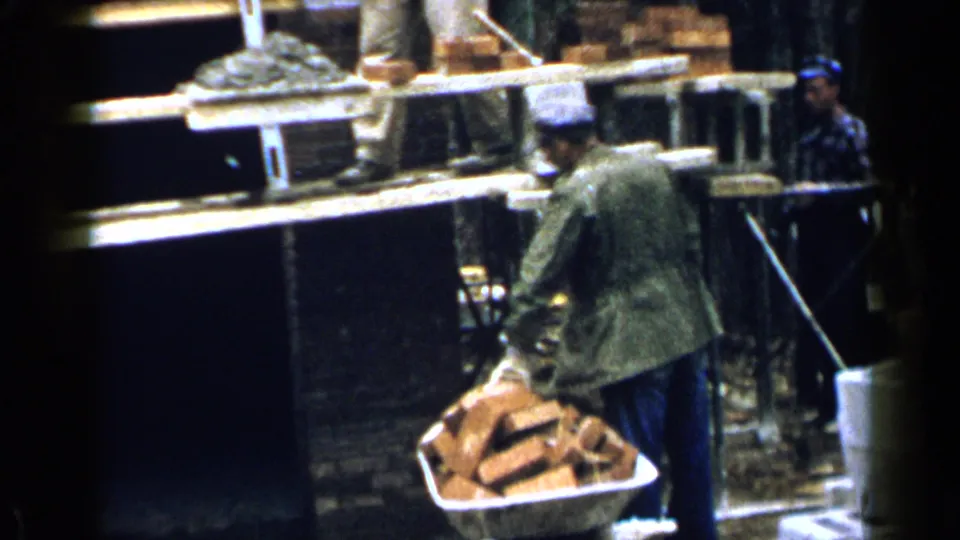 A worker in cap and jacket stands below house scaffolding with a wheelbarrow loaded with bricks in Birmingham, Alabama.